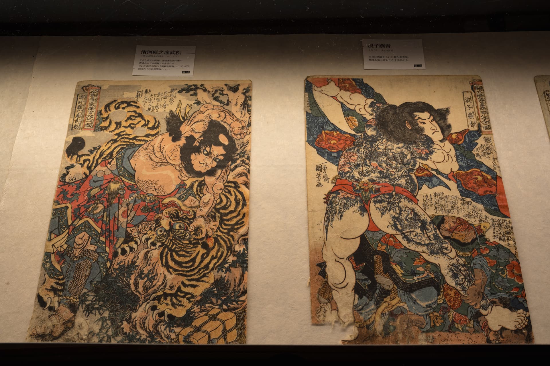 Two framed warrior prints side by side: a tattooed fighter wrestling a tiger, and a tattooed hero in blue and red robes