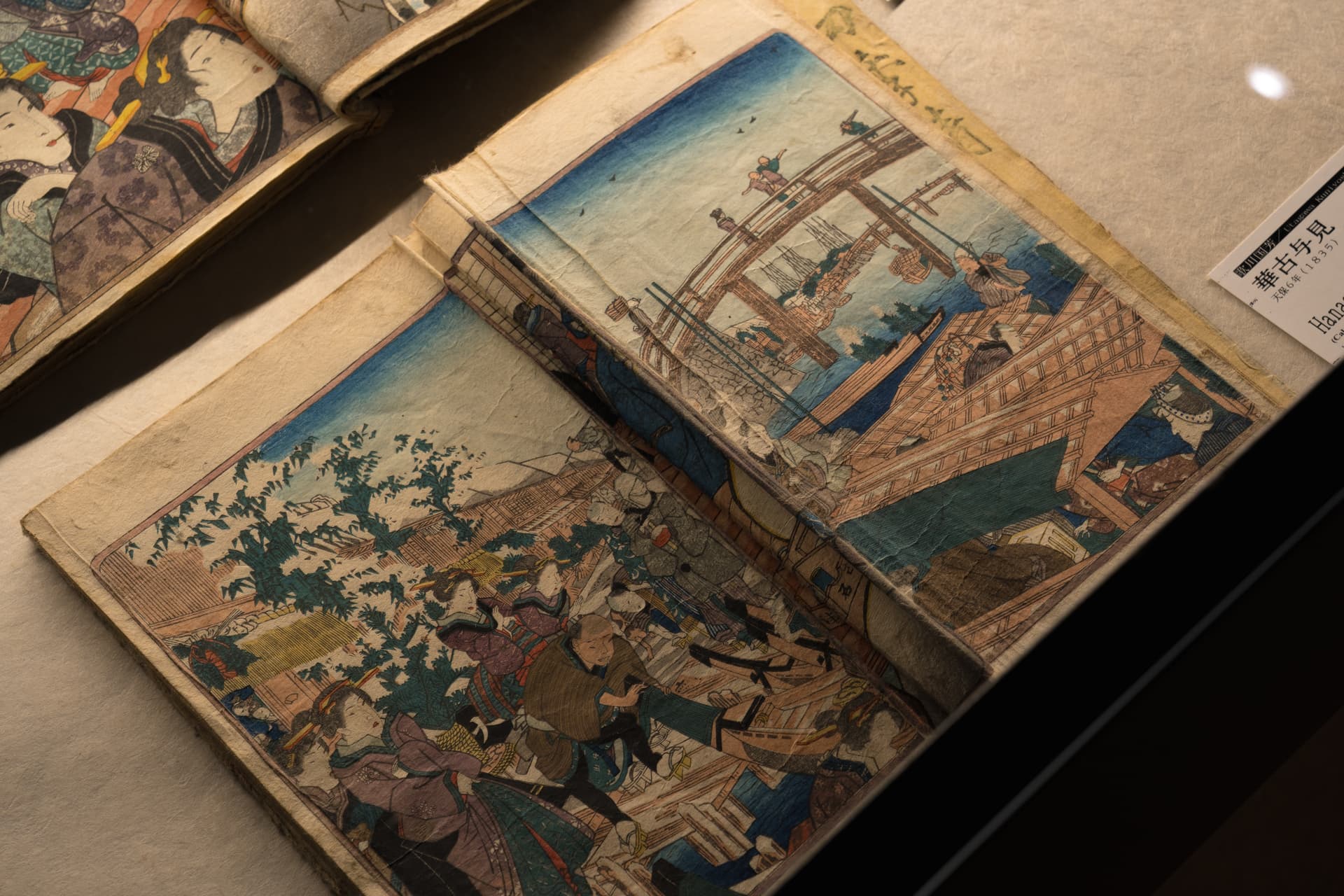 An open illustrated book in a display case, one page showing a crowded street below and the other a wooden bridge over a river