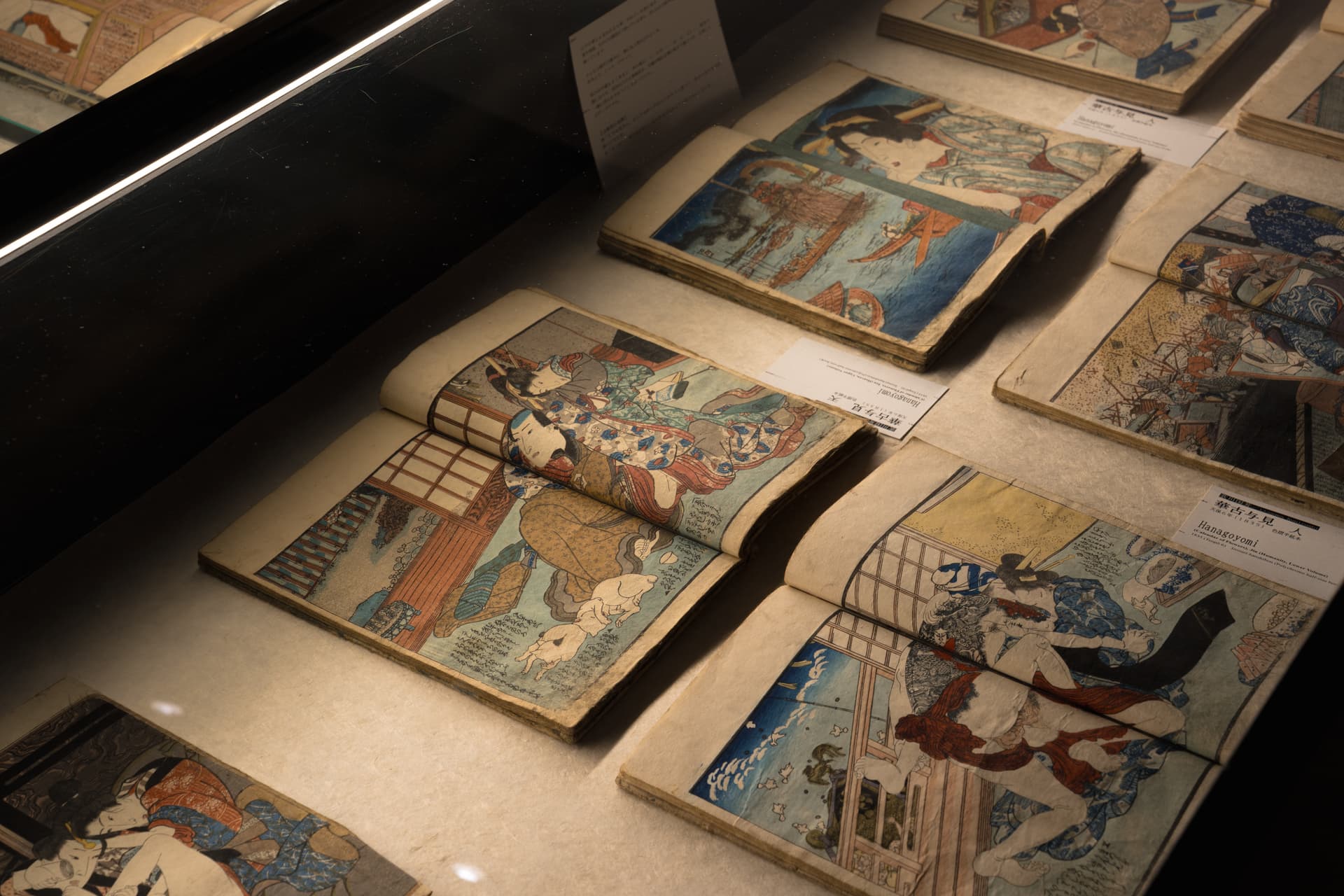 Several small Edo-period illustrated books lying open in a glass case, their colour pages showing couples in interiors