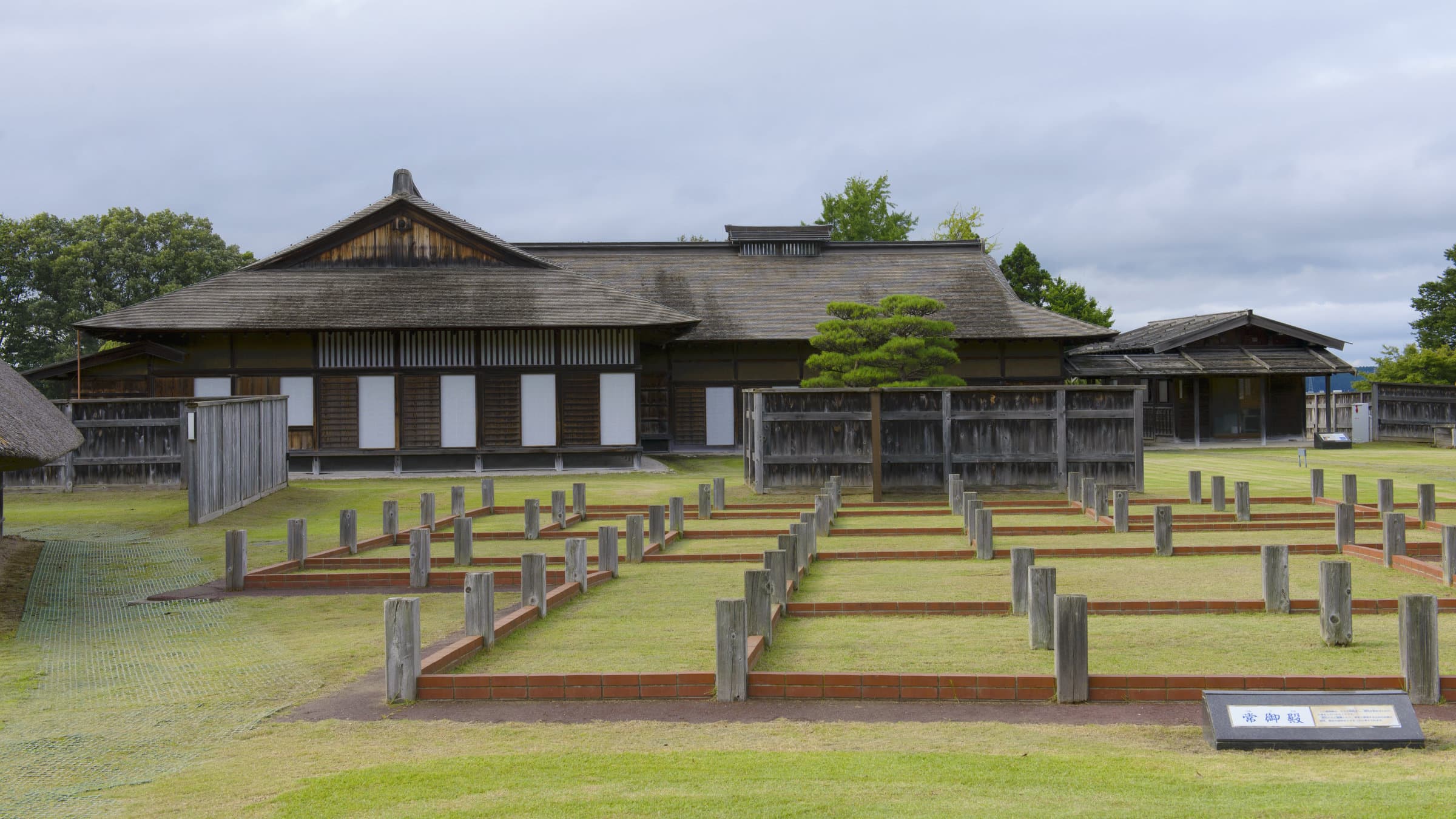 This image shows the reconstructed Nejo Castle site in Hachinohe, featuring traditional Japanese buildings with thatched roofs and wooden architecture. In the foreground, wooden foundation markers indicate where historical structures once stood, creating a grid pattern across the grassy grounds. The main building displays classic Japanese design elements including vertical wooden slats and white sliding panels. The archaeological park preserves the historical layout of this important castle site in Aomori Prefecture.