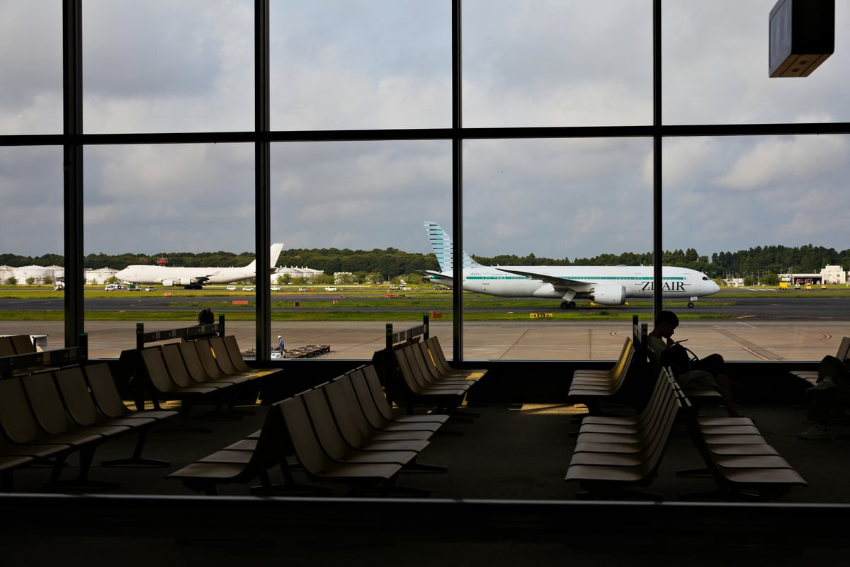 ZipAir Boeing 787 aircraft with teal livery parked on airport tarmac, viewed through terminal windows with passenger seating in foreground.