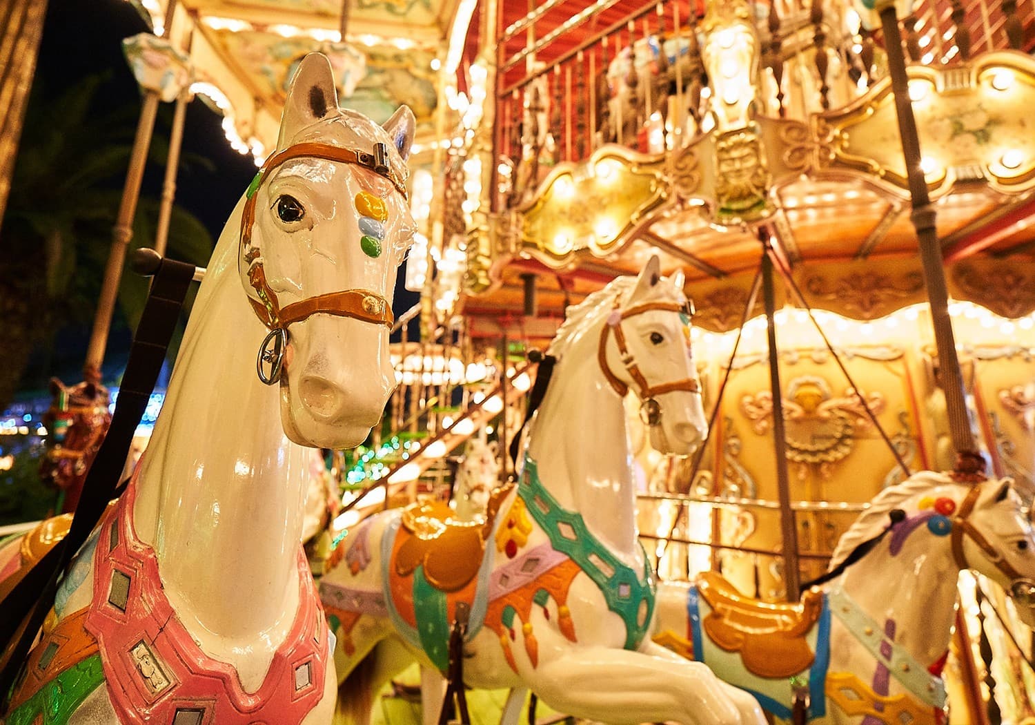 Carved white carousel horses with painted saddles under rows of warm lights