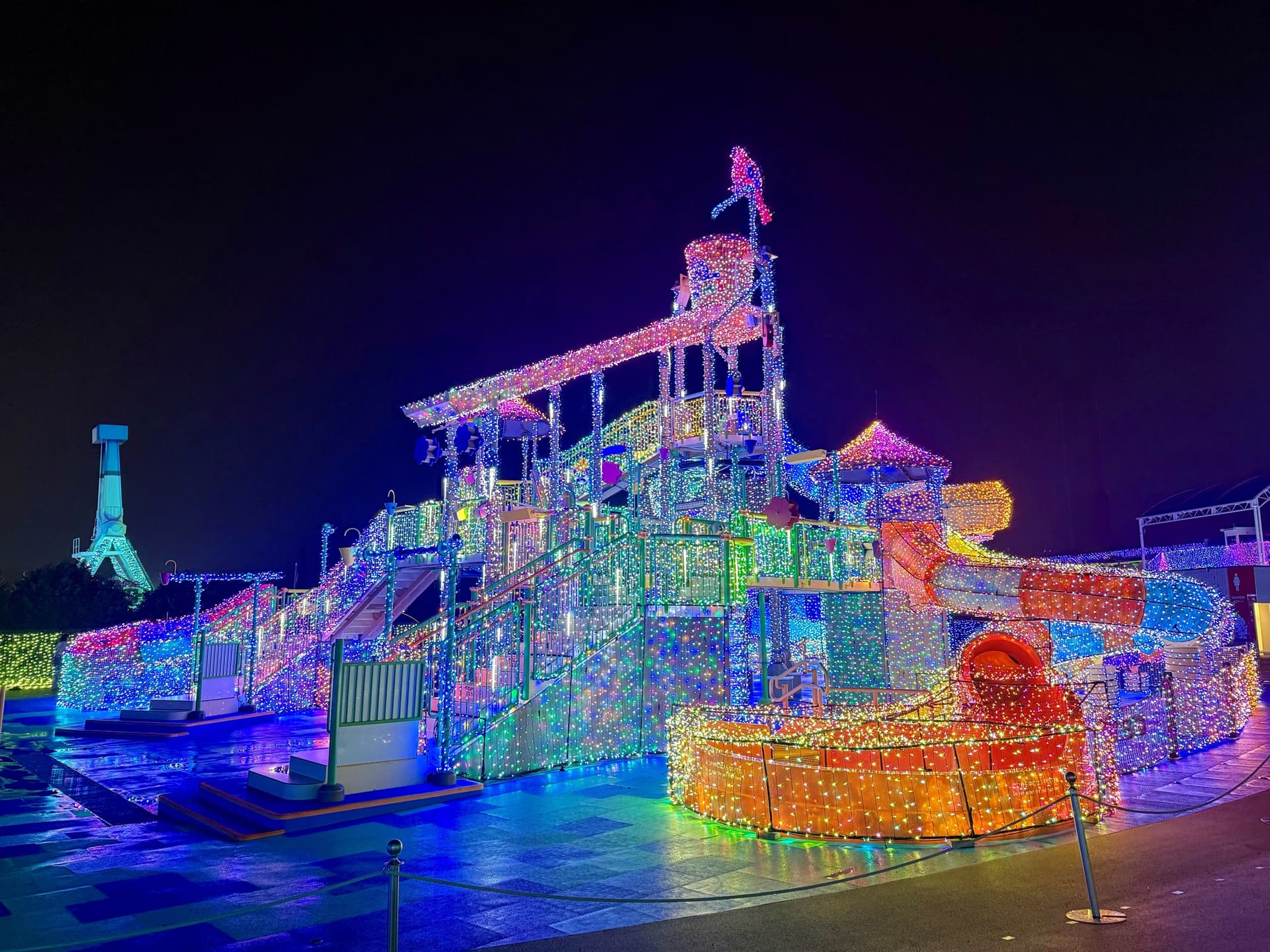 A large play structure with slides and towers covered in rainbow-coloured lights at night
