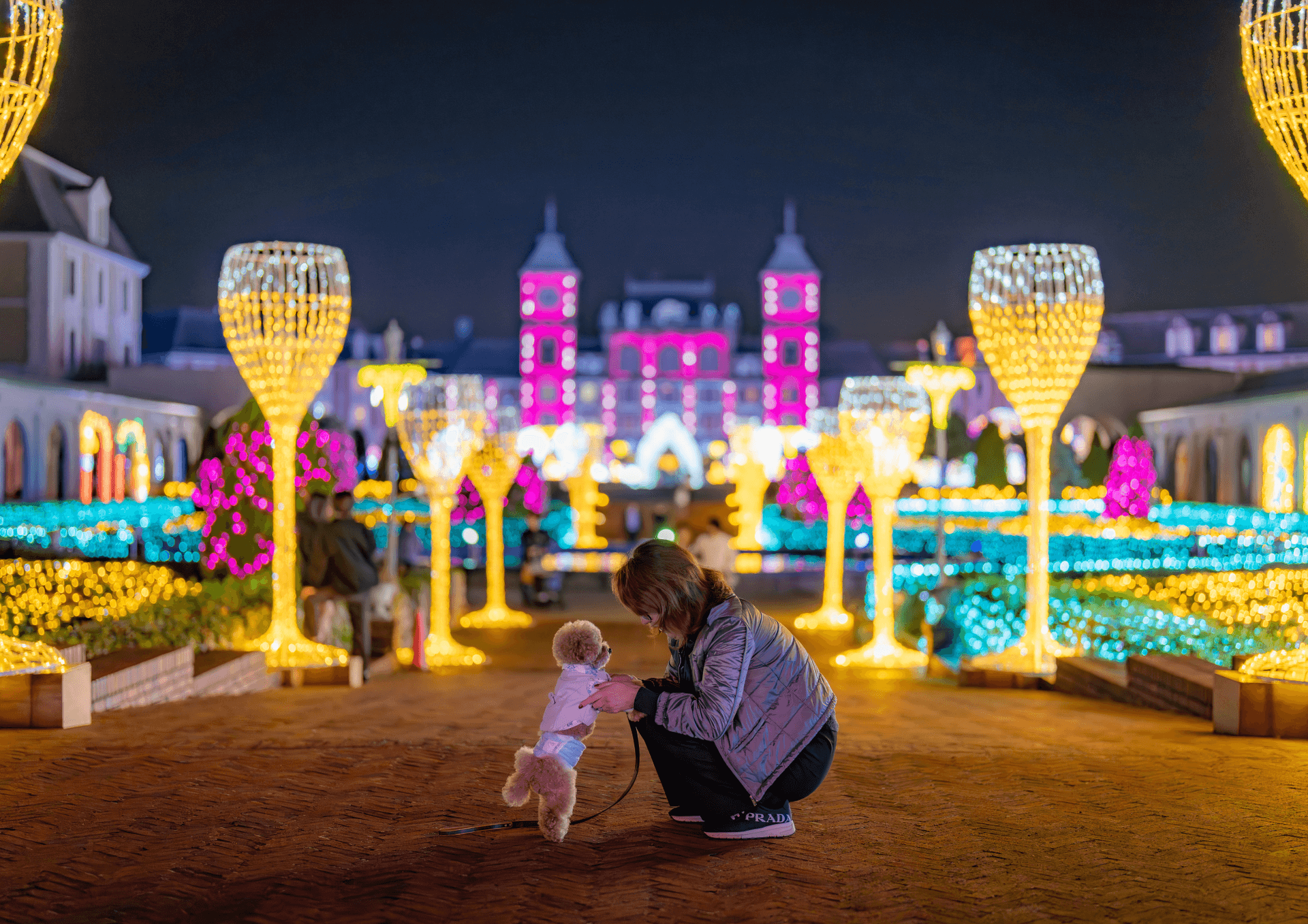 A pink-lit palace at the end of an avenue of tall glowing lights shaped like wine glasses, with a woman and a small dog in the foreground