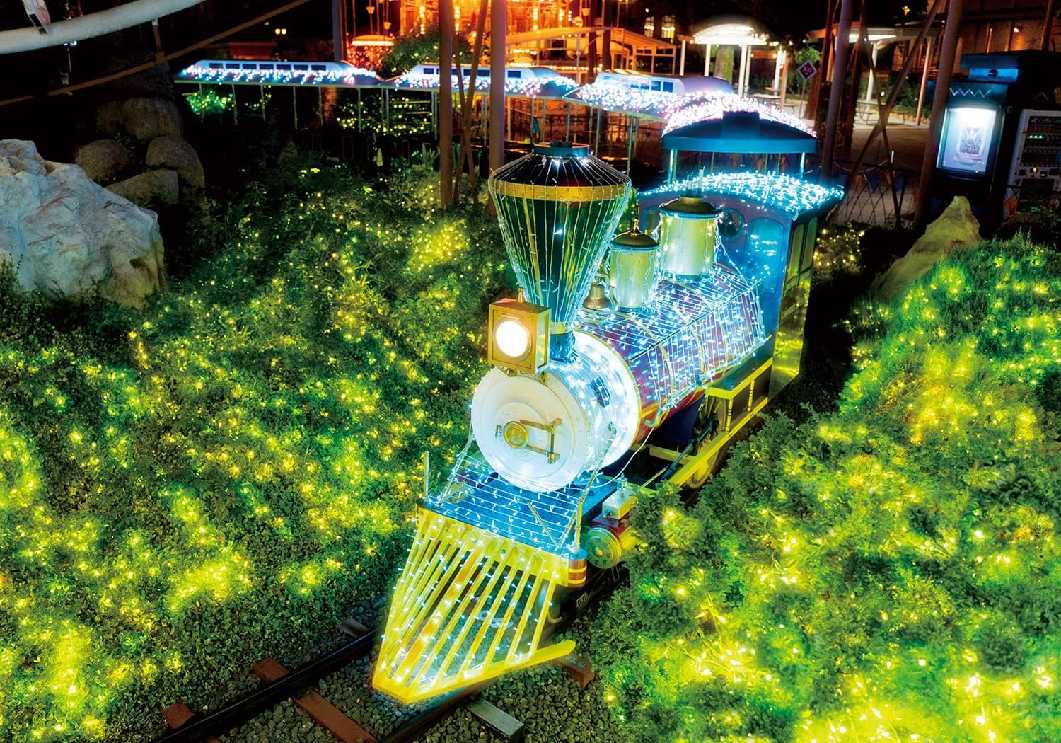 A small locomotive outlined in blue and white lights on a track through bushes covered in green lights