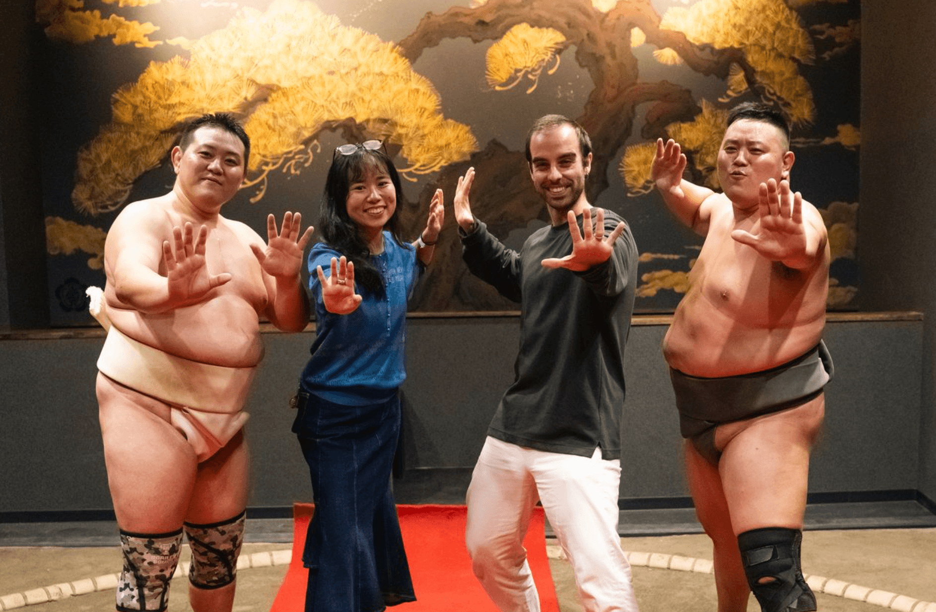 Two guests posing with two sumo wrestlers in the ring, in front of a painted pine tree