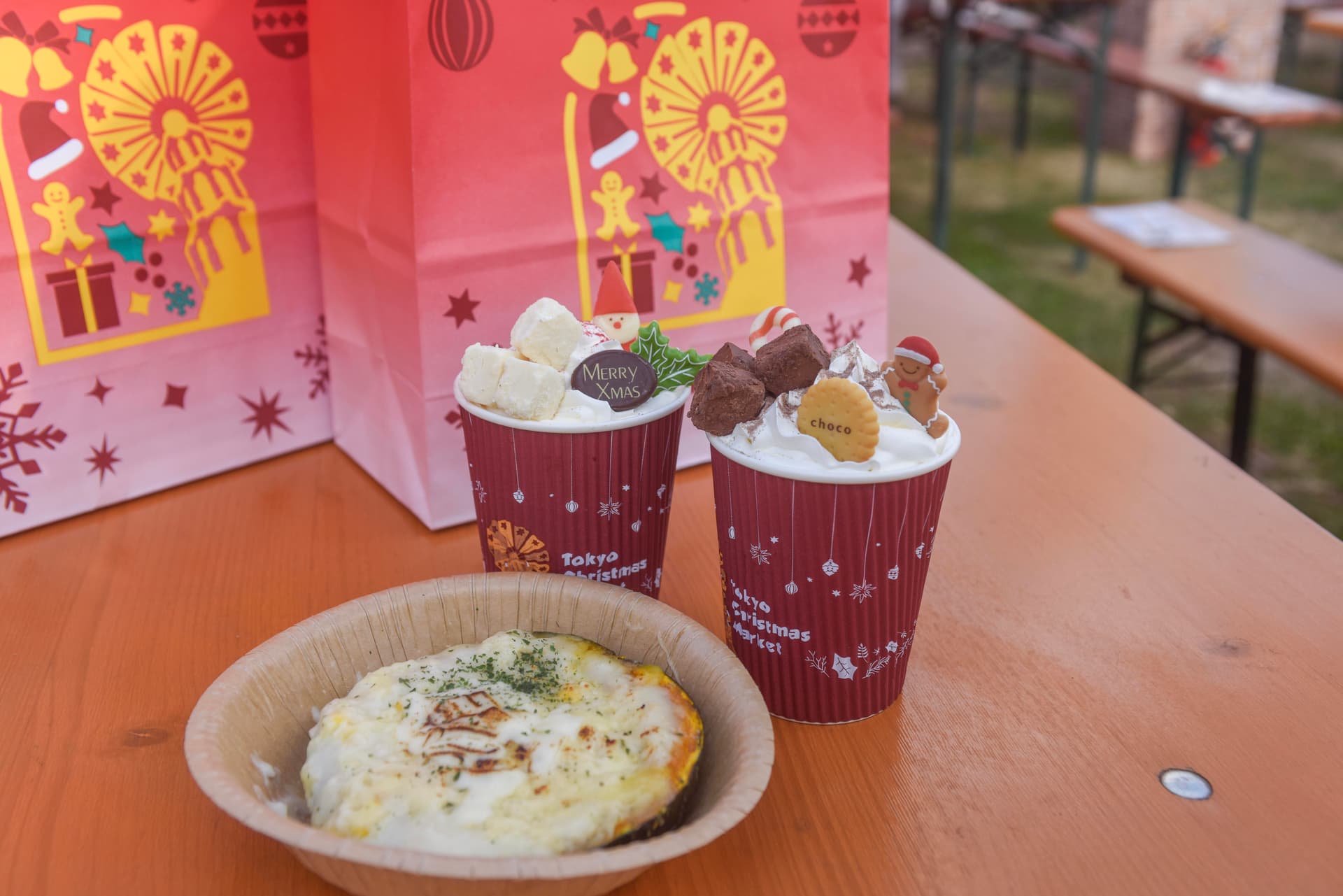 Two hot drinks in red Tokyo Christmas Market cups, topped with cream, marshmallows and Christmas sweets, beside a cheese-topped bake on a wooden table