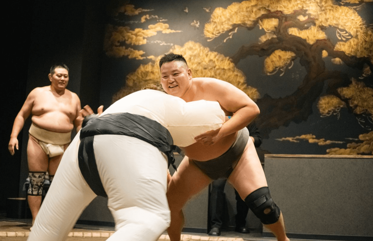 A guest in a padded white sumo suit pushing against a wrestler in the ring