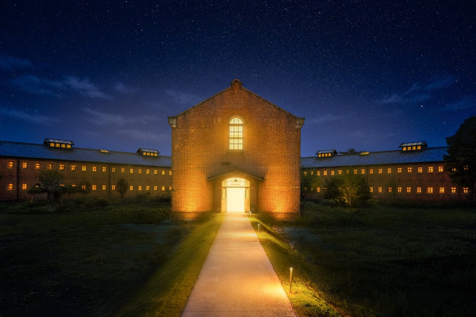 The red-brick central building of the former Nara Prison lit under a starry sky, with a lit path leading to its door