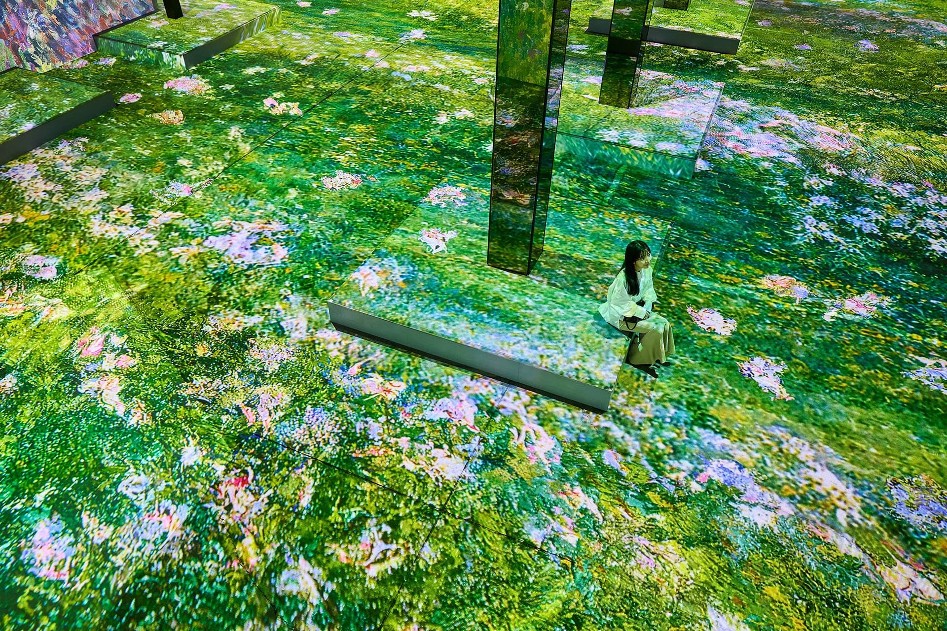 A visitor sitting on a floor covered in projected flowers and greenery, with mirrored pillars rising from it
