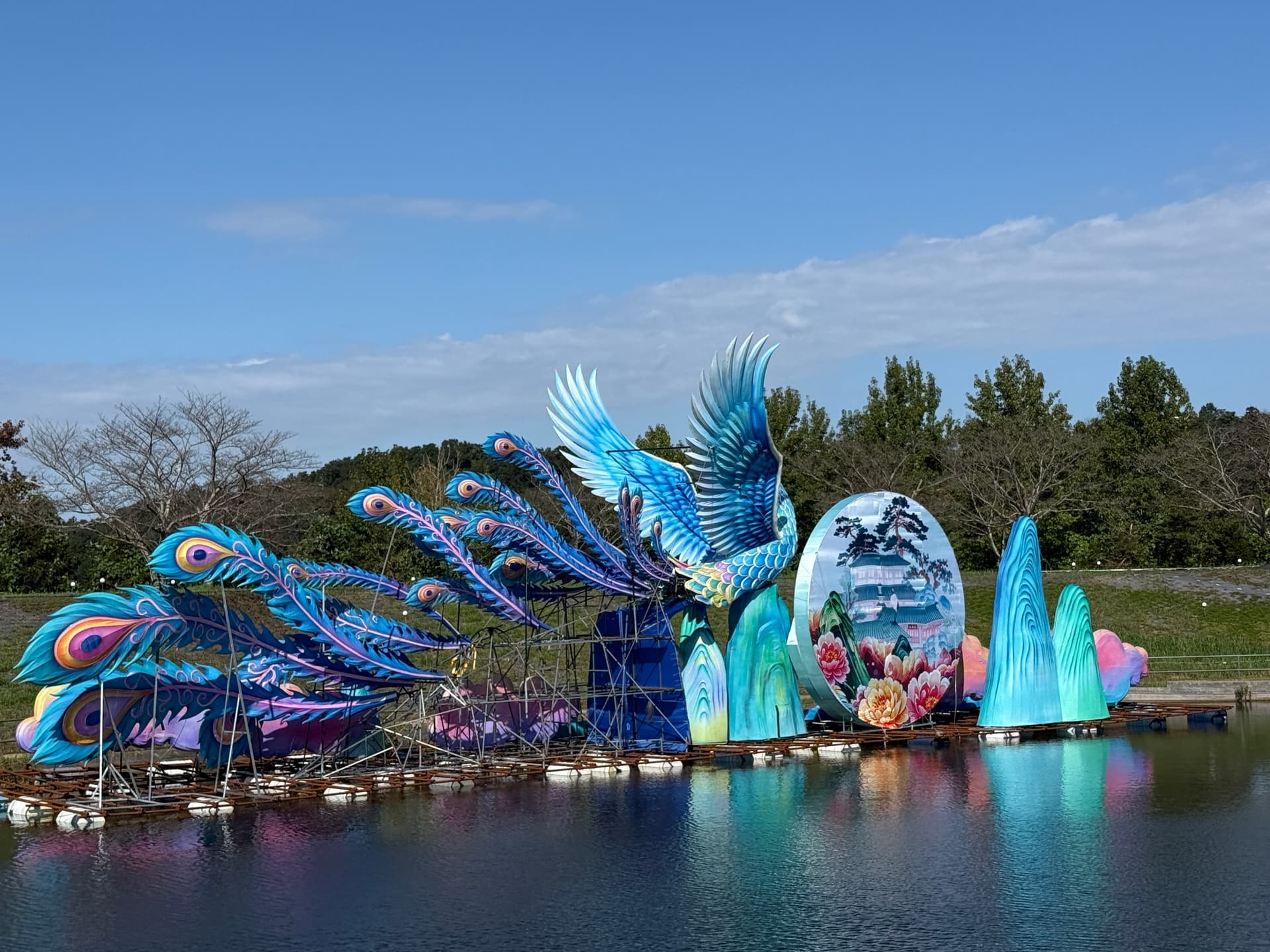 The peacock lantern being built on the lake at Tokyo German Village in 2026, beside a round painted panel and blue rock shapes