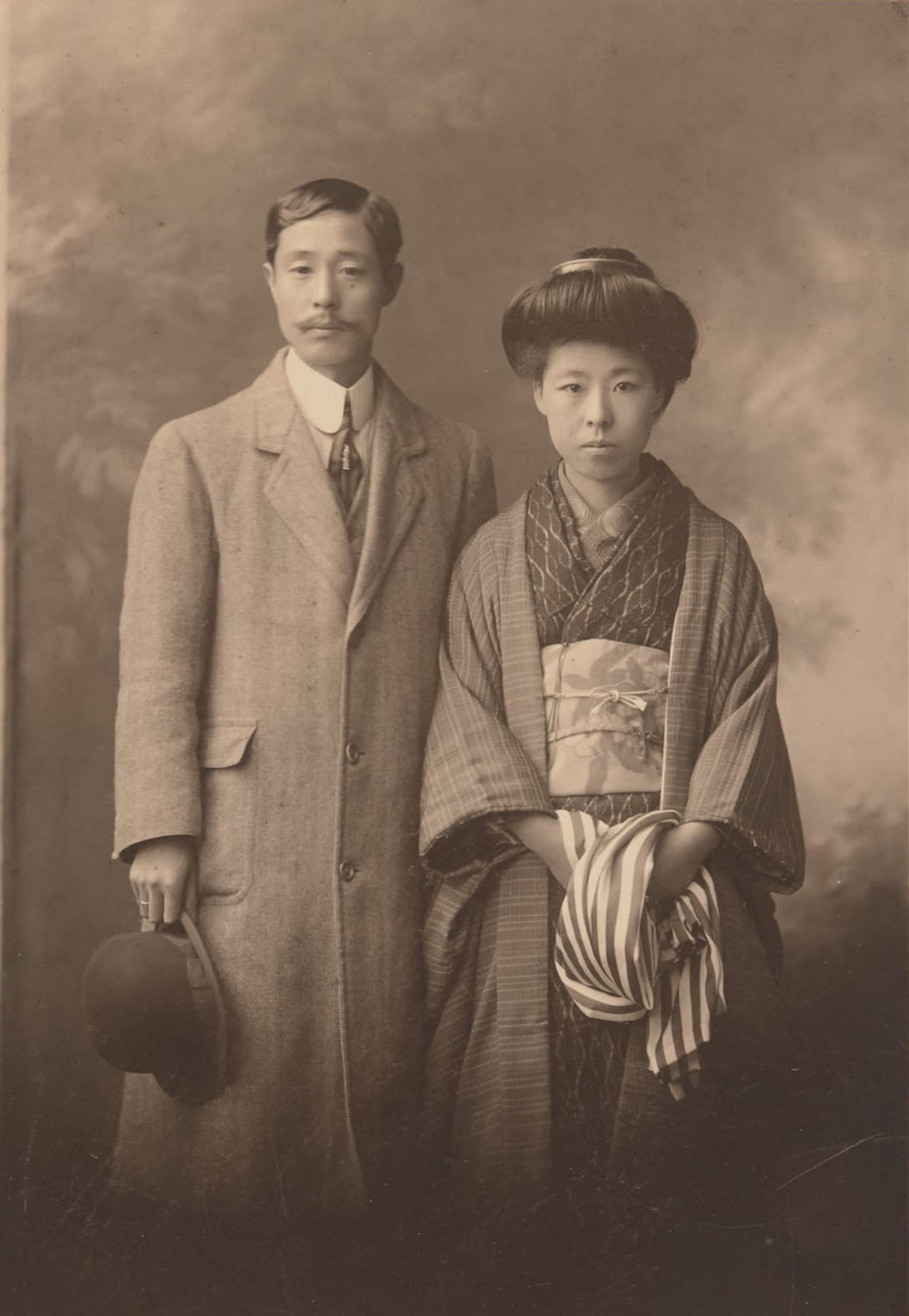 Sepia photograph of a man in a long coat holding a hat beside a woman in kimono with a shawl