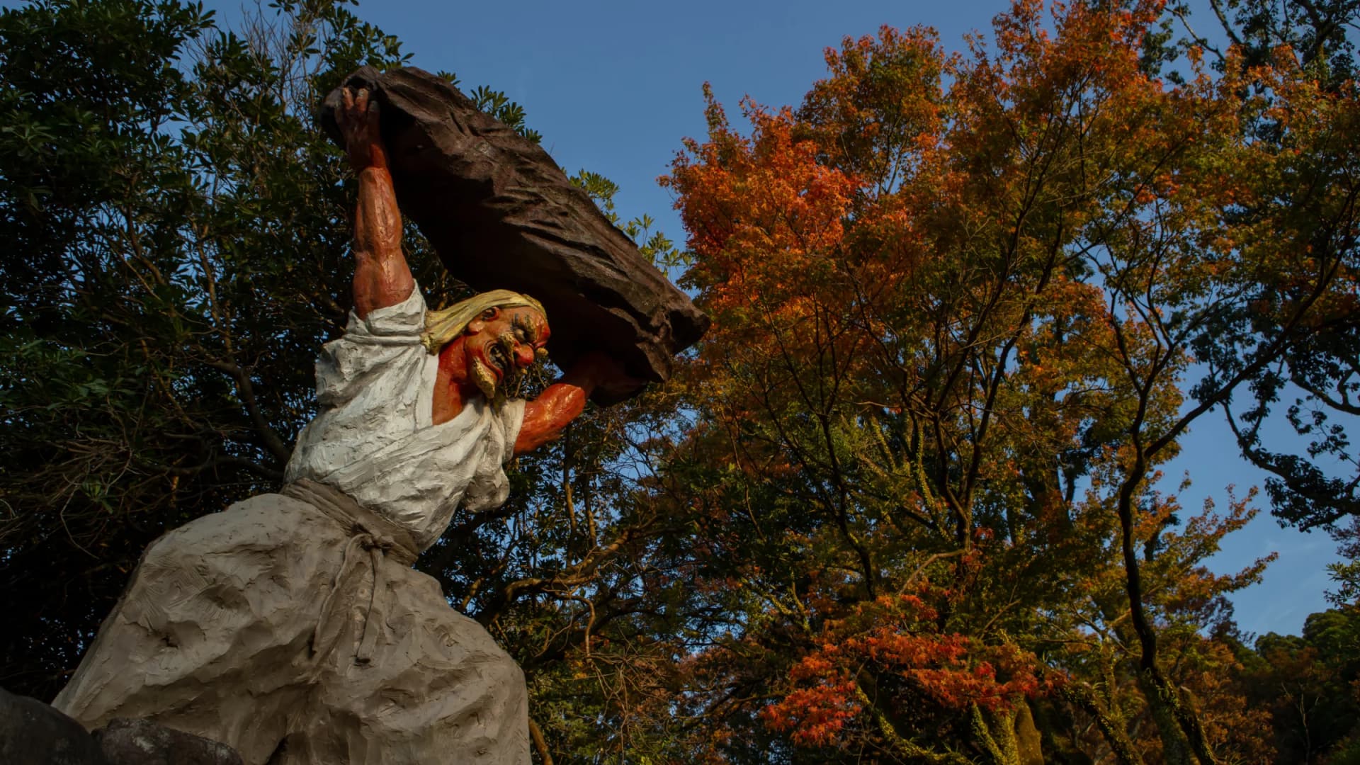 A statue of Ame-no-Tajikarao hurling open the heavenly rock cave door stands against autumn foliage at Amano-Iwato Shrine, Takachiho