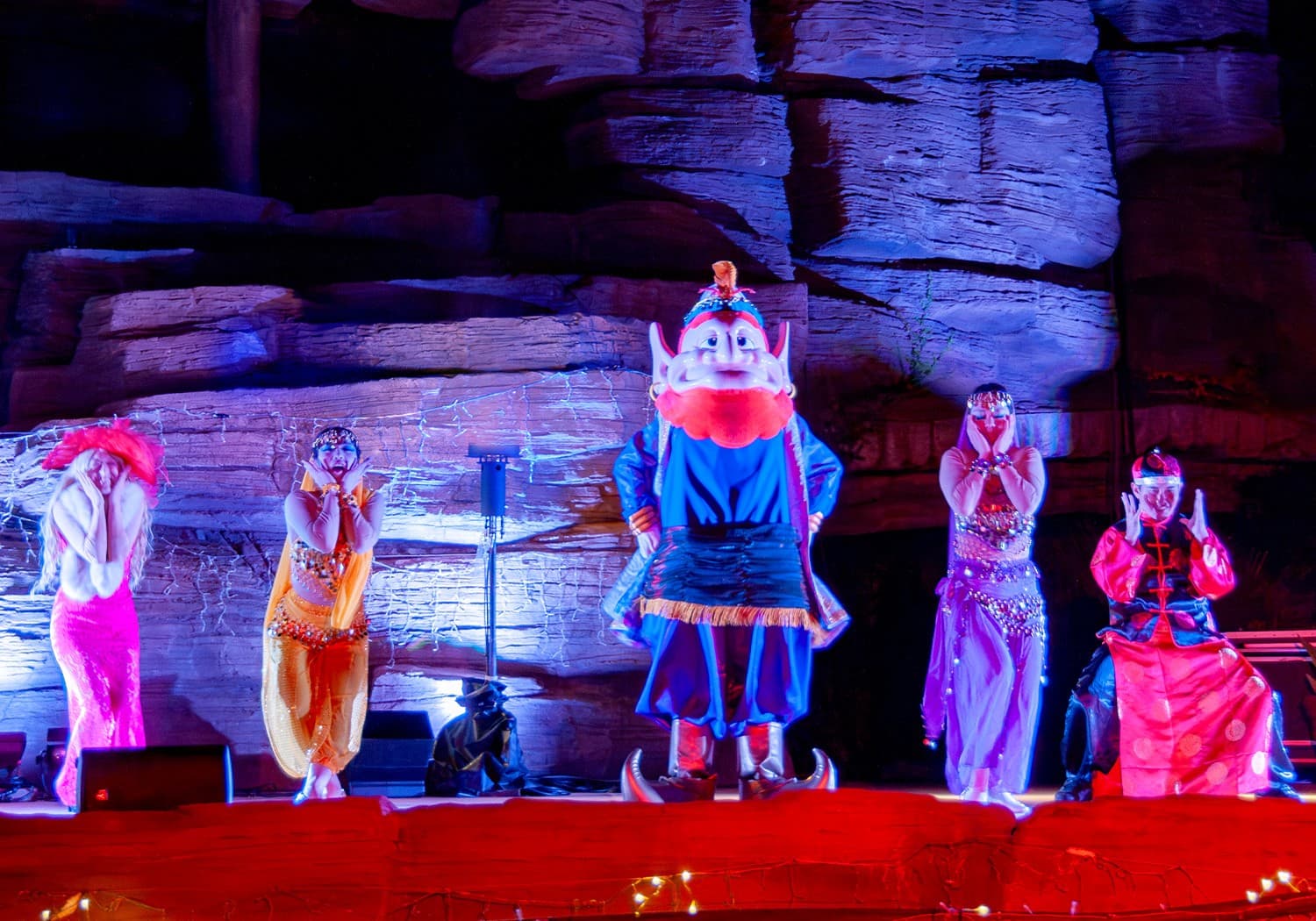 Costumed characters and dancers in bright robes on a stage lit blue and red in front of a rock wall