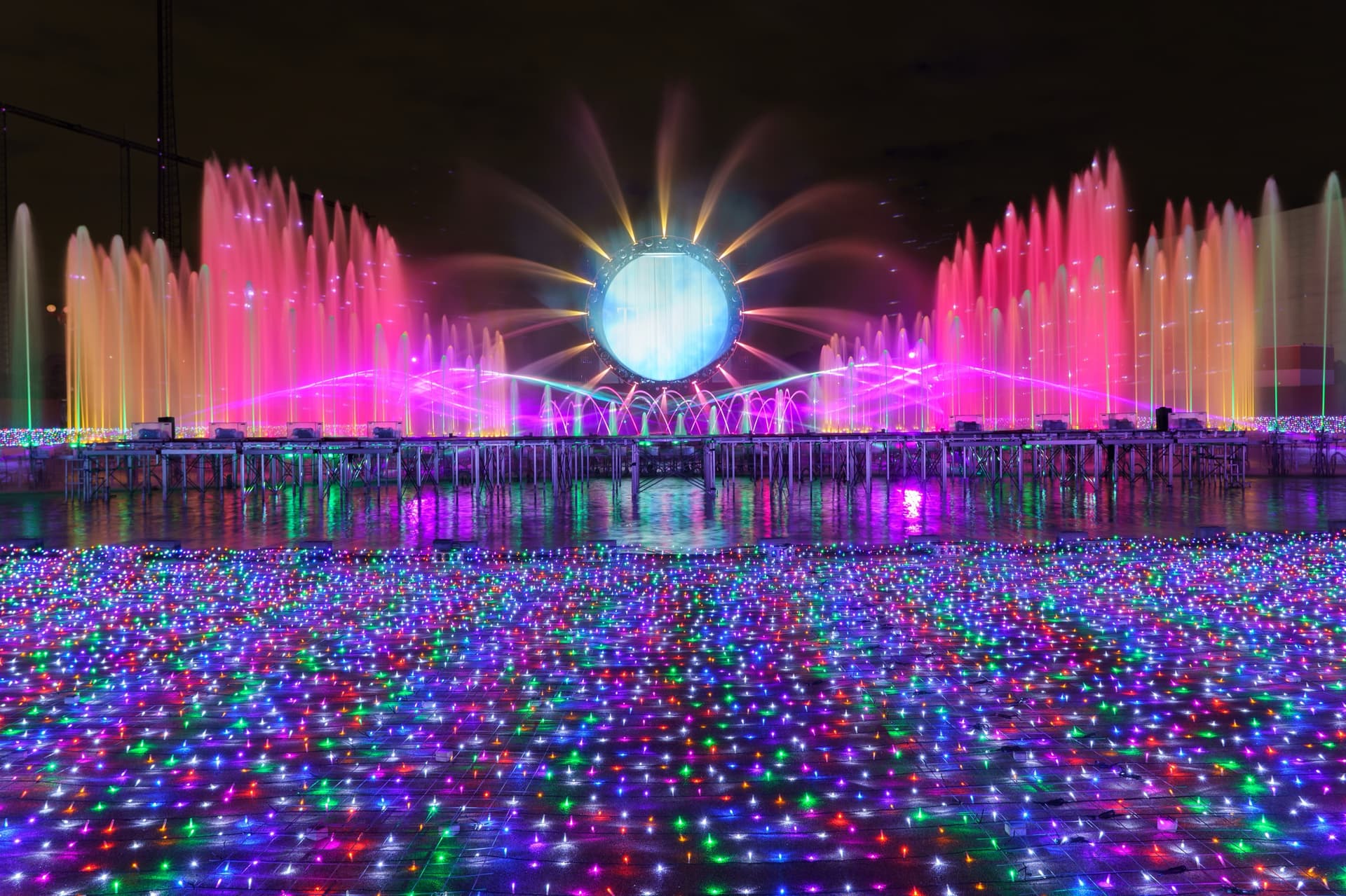 A glowing ring-shaped screen above rows of pink fountains, with a pool of coloured lights in front