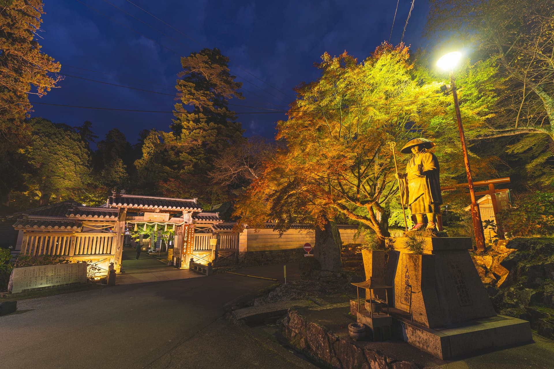 A tiled temple gate at dusk, a lit red and yellow maple, and a bronze statue of a monk with a hat and staff on a stone base