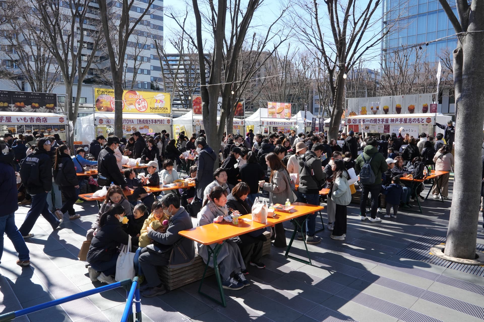 Crowds eating at long wooden tables under bare trees, with white food stalls behind them