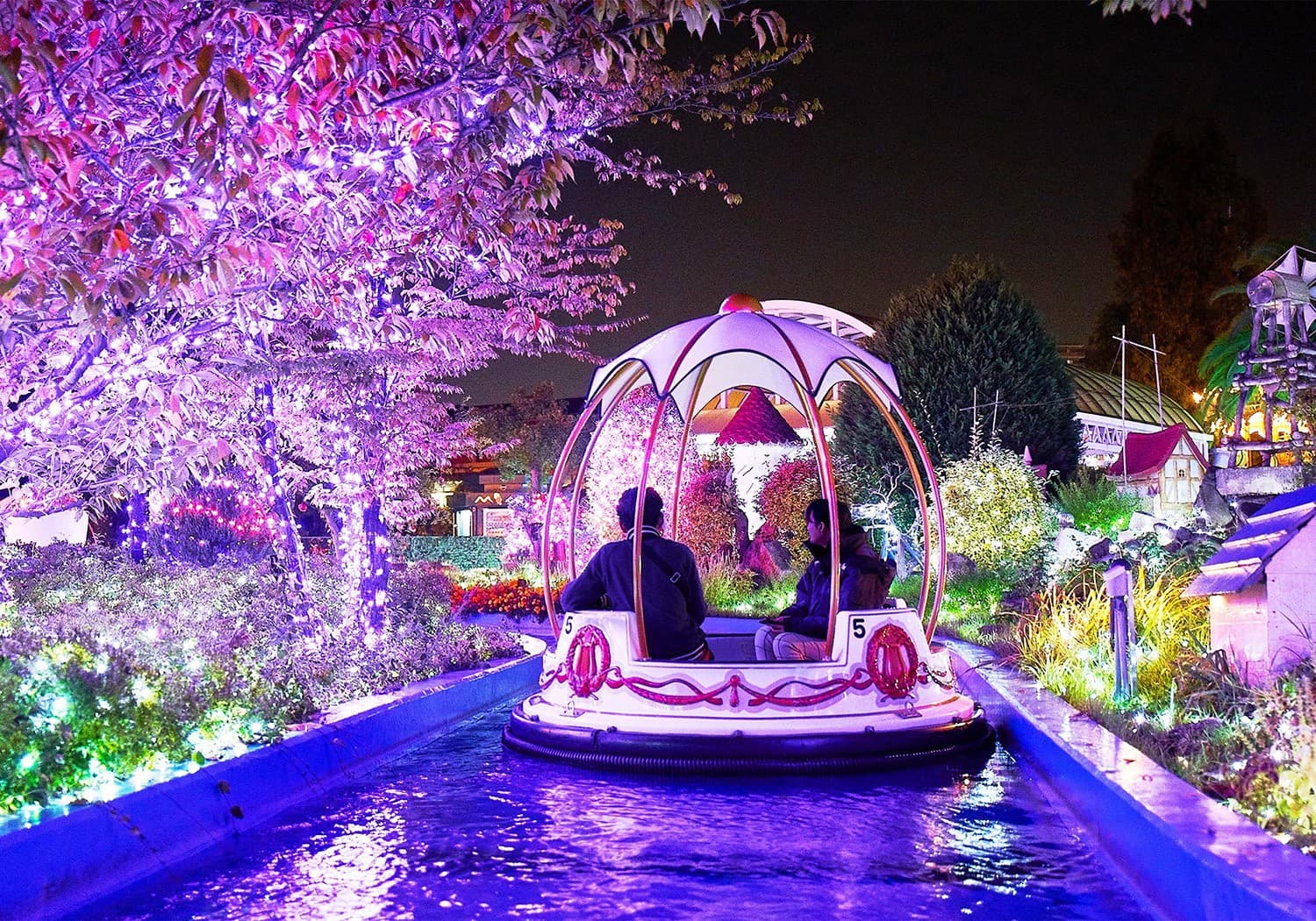 A domed boat with two riders on a narrow waterway lit purple, under trees covered in pink lights