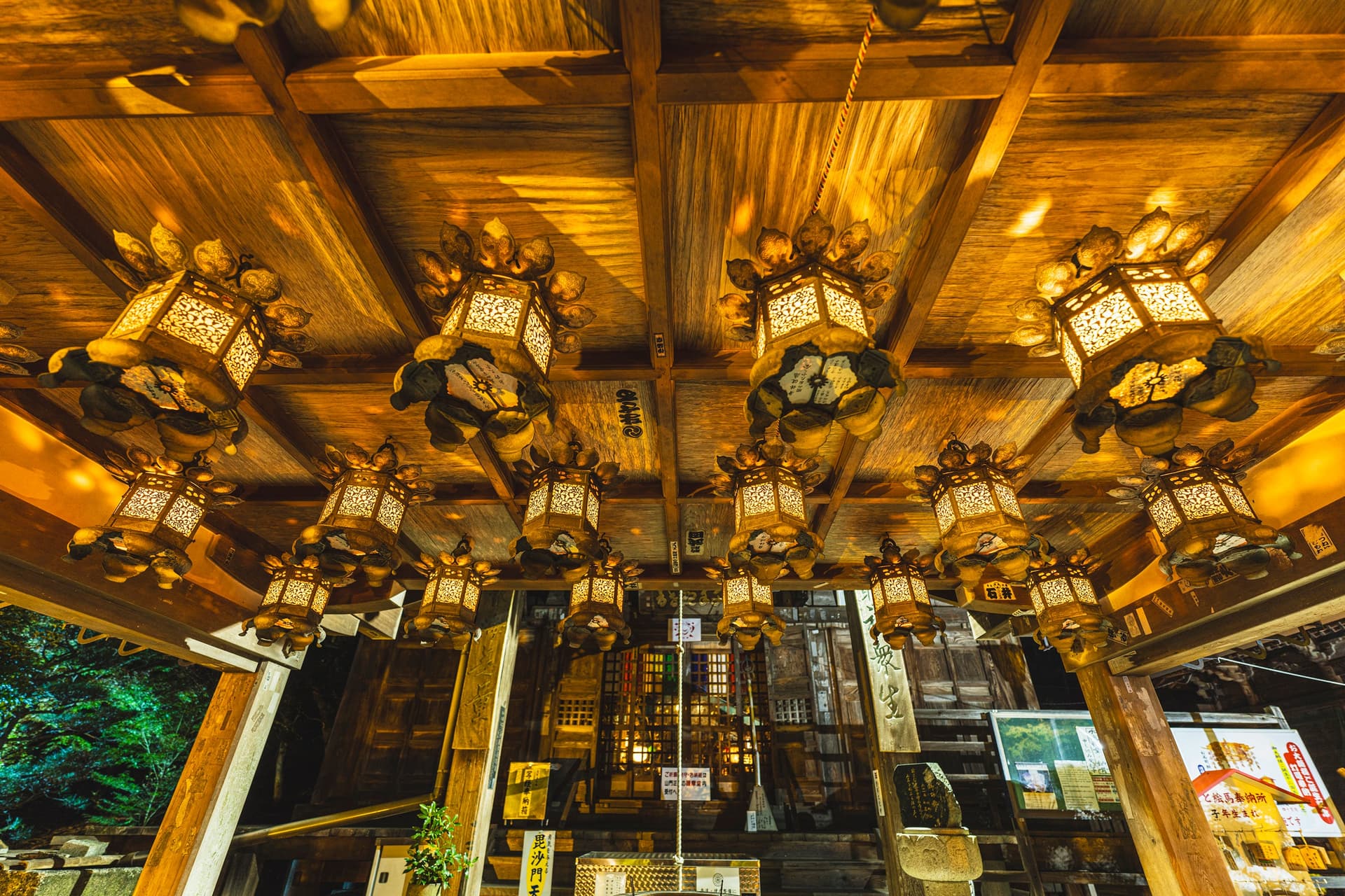 Rows of ornate bronze lanterns lit gold under the wooden ceiling of a temple porch, the hall's lattice doors behind