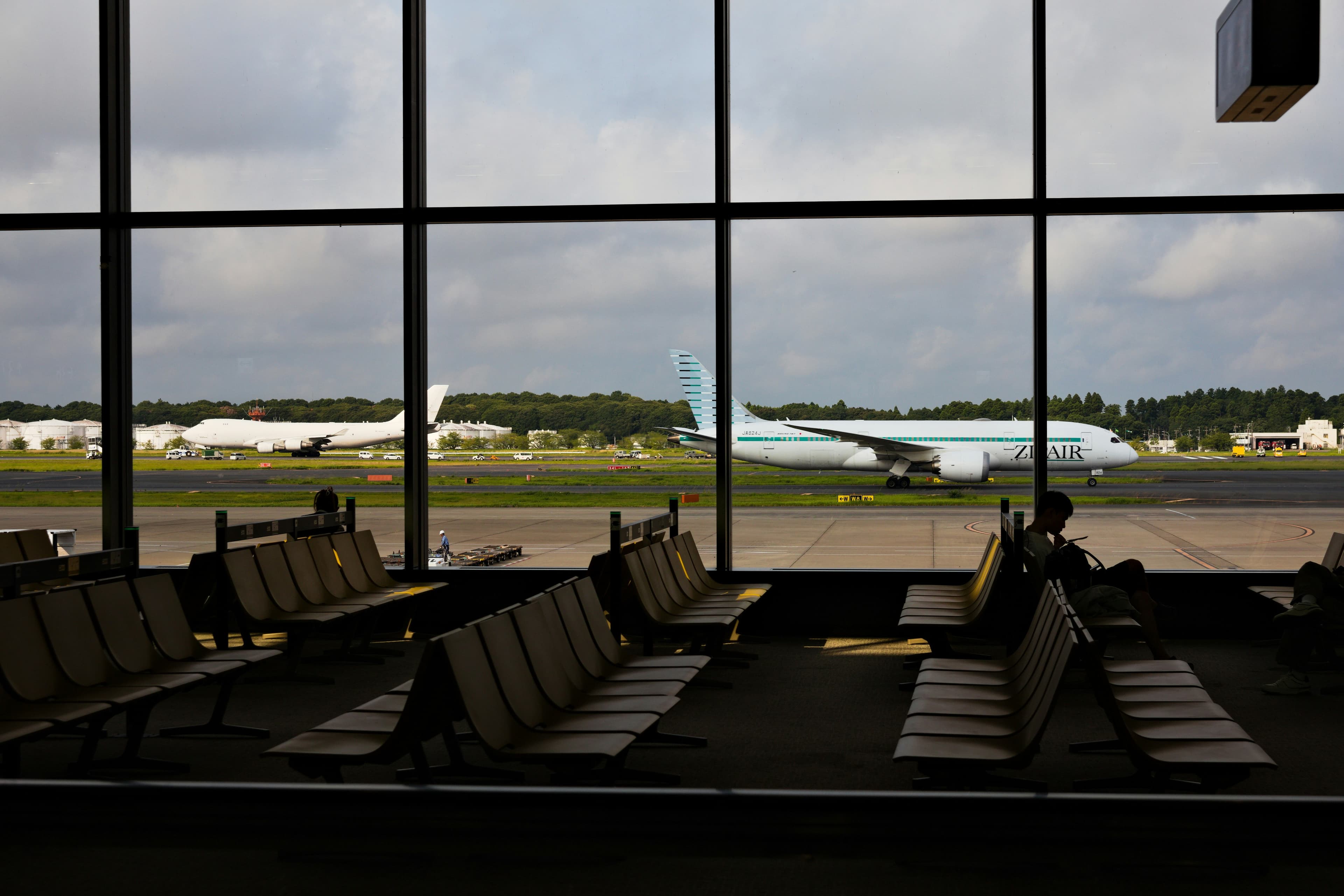 ZipAir Boeing 787 aircraft with teal livery parked on airport tarmac, viewed through terminal windows with passenger seating in foreground.